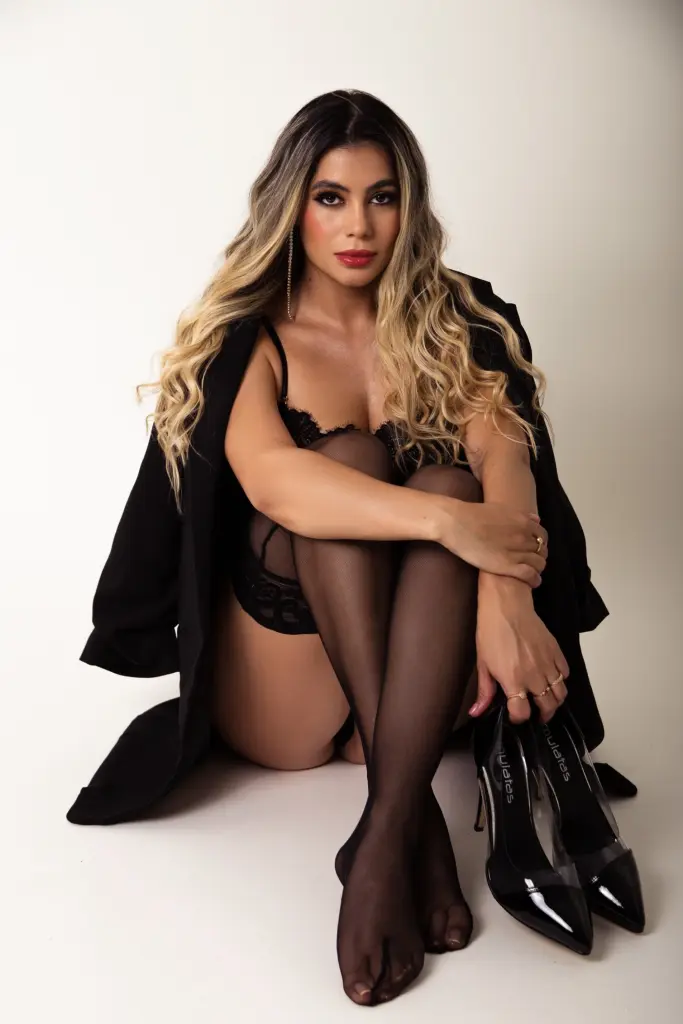 Dapne_Rogers poses sideways against a wall, wearing a tight black long-sleeve top that shows her curves and long blonde hair.
