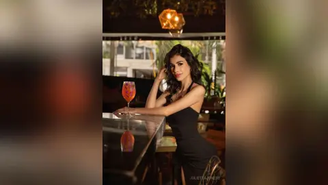 Elegant woman in a black cocktail dress holding a drink at a moody, dimly lit upscale bar.