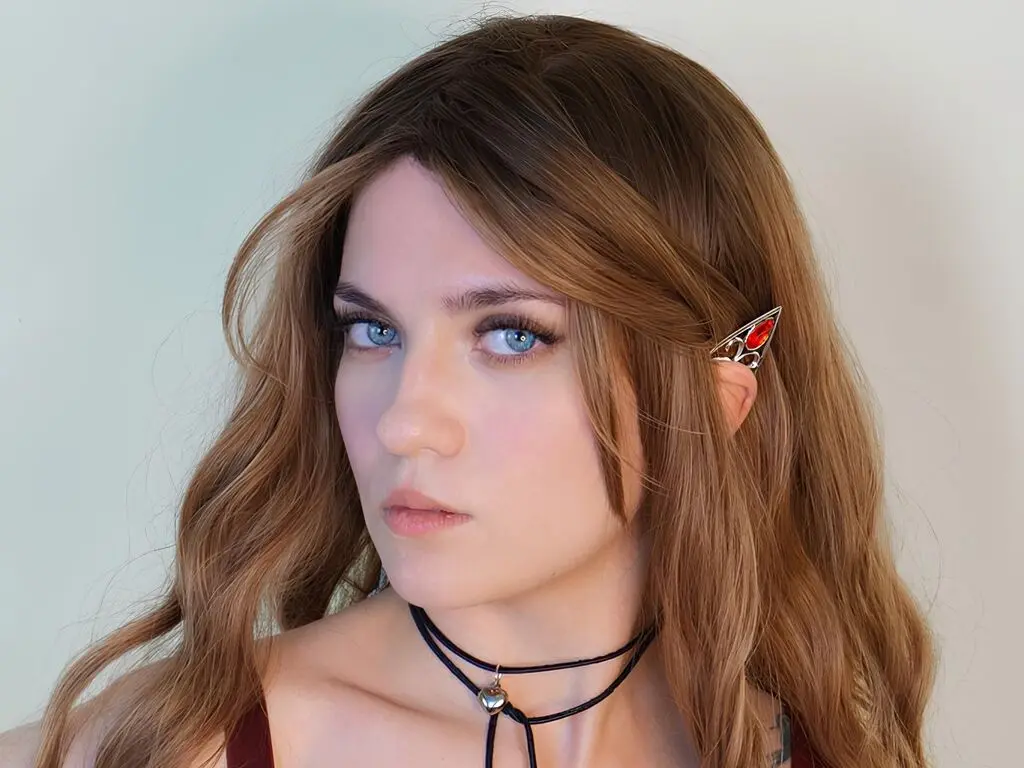 Young woman with wavy brown hair and a black choker posing against a plain background with a calm expression.