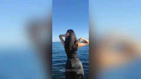 Woman in a sleek black dress standing by the ocean under a clear blue sky with a serene mood.