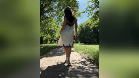 CassyTorres walks away down a sunlit path wearing a short, white slip dress with black lace trim and white high heels.