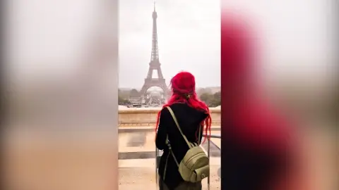 Woman with red hair in a black outfit standing before the Eiffel Tower in Paris on a cloudy day.