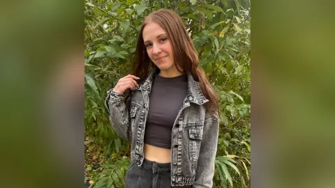 Smiling young woman in a grey denim jacket and black top posing in a lush green forest setting.