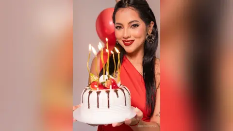 Smiling woman in a red dress holding a white birthday cake with lit candles against a festive red background.