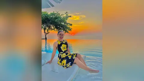 Smiling woman in a yellow floral dress sitting by a pool during a golden sunset at a tropical resort.