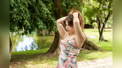Young woman in a floral summer dress posing thoughtfully in a lush green park by a peaceful lake.