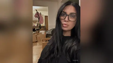 Young woman with long dark hair and glasses wearing a black turtleneck in a cozy indoor setting.
