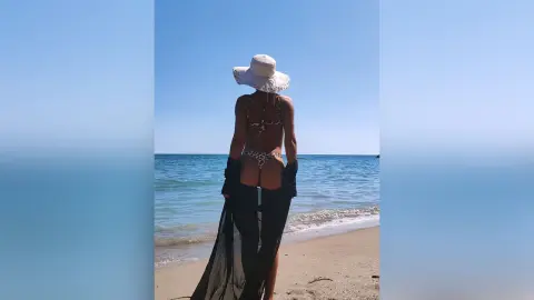 Woman in a white sun hat and leopard print bikini standing on a sunny beach facing the blue ocean.