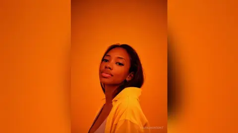 Young woman in a yellow shirt posing against a warm orange background with a soft, moody aesthetic.