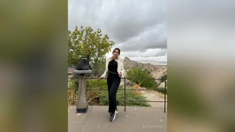 AlanaPearce stands on a balcony wearing a white jacket and black pants, posing against a cloudy sky and a rocky landscape.