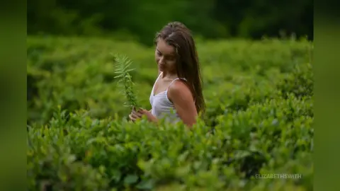 Young woman in a white tank top holding a green fern in a lush, peaceful green field.