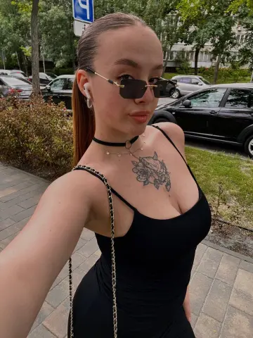 Young woman in a black tank top and sunglasses with a floral chest tattoo standing outdoors on a sunny day.