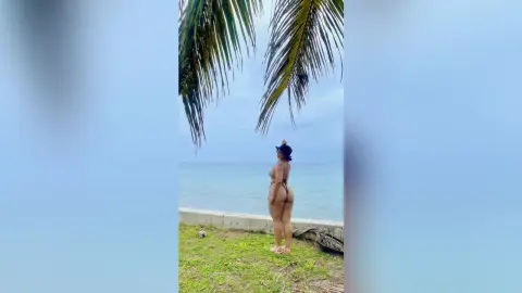Curvy woman in a black bikini standing on a tropical beach under palm trees on a sunny day.