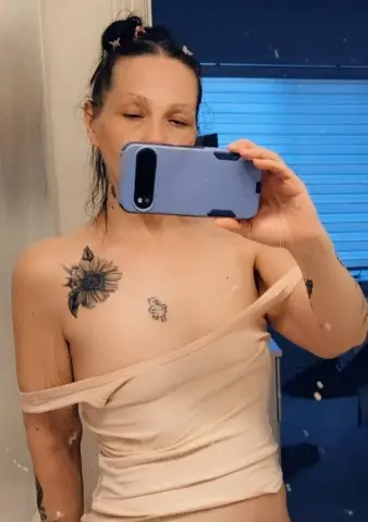 Woman with sunflower tattoo in beige tank top taking a mirror selfie in a dimly lit room.