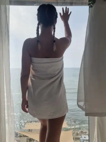 Woman in a white towel looking out a window at the ocean view during a relaxing seaside vacation.