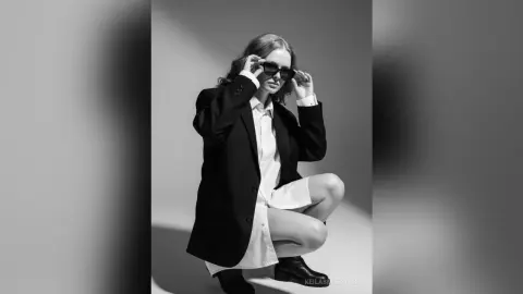 Stylish woman in a black blazer and white shirt wearing sunglasses in a minimalist black and white studio setting.