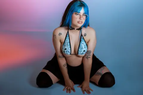 Blue-haired woman in a patterned bikini and black skirt posing confidently against a colorful studio background.