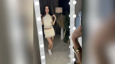 Young woman in a cream lace crop top and mini skirt posing in a lighted vanity mirror for a chic aesthetic look.