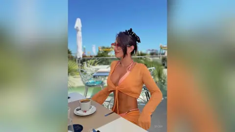 Stylish woman in an orange crop top and sunglasses enjoying coffee at a sunny outdoor cafe terrace.