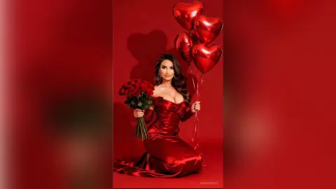 AnnaKarev poses in a red dress holding a bouquet of red roses against a red background with heart balloons.