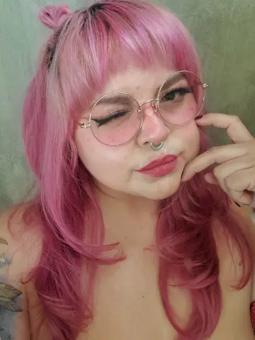 Smiling woman with pink hair and round glasses posing in a casual indoor setting.
