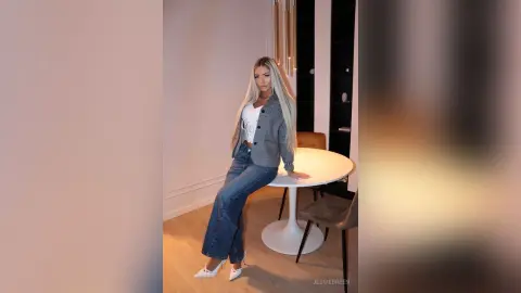 Blonde woman in grey blazer and wide-leg jeans posing by a white table in a modern minimalist room.