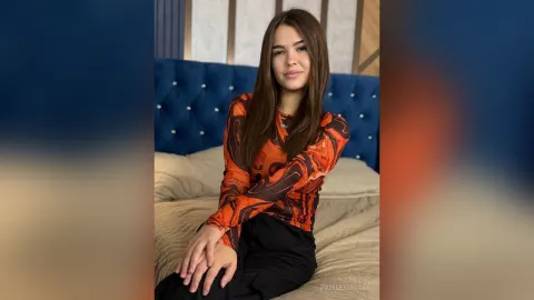 PaisleyMoore — PaisleyMoore sits on a bed wearing an orange patterned long-sleeve top and black pants.