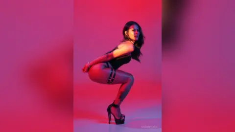 Glamorous woman in black bodysuit and high heels posing against a vibrant pink and red studio background.