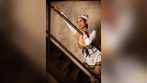 JuliahLewis poses leaning against a dark banister wearing a white and blue sailor-style outfit with a red necktie.