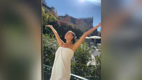 AnaMathers stands on a balcony with her arms raised, wrapped in a white towel against a sunny outdoor background.