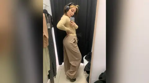 Woman in beige lounge set and headphones taking a mirror selfie in a dressing room.