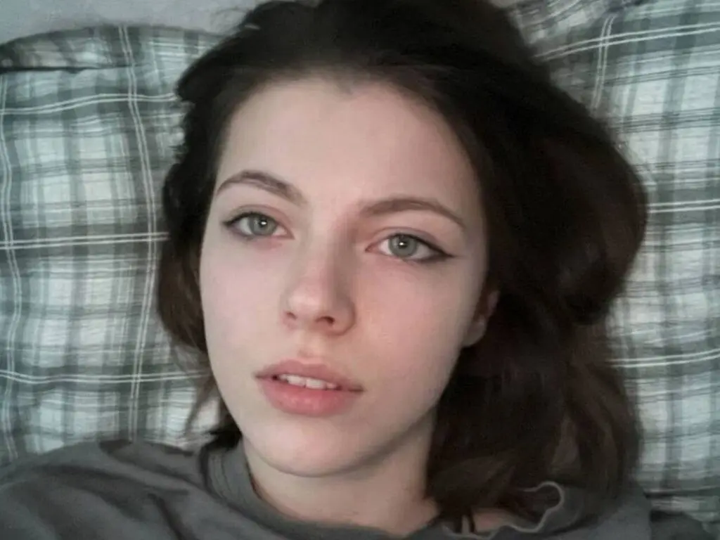 Young woman with brown hair and winged eyeliner relaxing on a plaid bedspread in a moody, casual setting.
