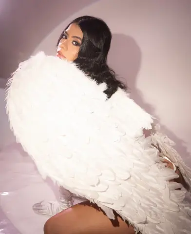 Beautiful woman in a white feathered angel costume posing against a soft pink background.