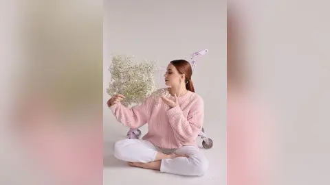 Young woman in a pink knit sweater sitting cross-legged with baby's breath flowers in a dreamy aesthetic setting.