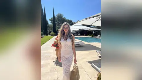 LilyReyes stands outdoors in a tight, white mesh dress, showing her long blonde hair and glasses while standing next to a swimming pool.