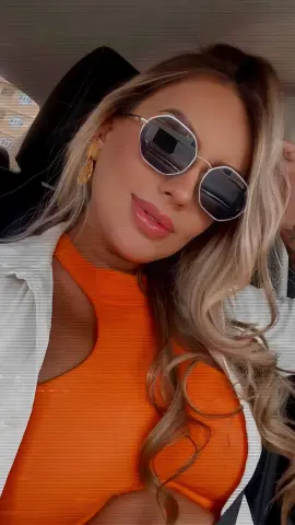 Blonde woman in orange top and white shirt wearing sunglasses sitting in a car with a relaxed summer mood.
