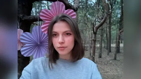 Young woman in a grey sweatshirt smiling amidst pink paper decorations in a tranquil forest setting.