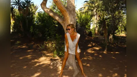 AtlamaDesmond wearing a white tank top, sitting against a large tree trunk in a sunny, wooded outdoor setting.