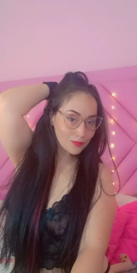 sussy31 — Young woman with long dark hair and glasses wearing black lace lingerie in a cozy pink bedroom.