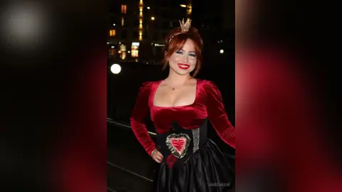 Smiling woman in a red velvet gown and crown posing at night in a glamorous evening setting.
