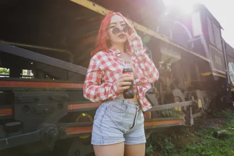 LiahCollinss poses outdoors in a red and white checkered shirt and denim shorts, holding a glass bottle of soda in front of a large train.