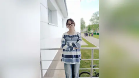 Young woman in a striped long-sleeve shirt and jeans standing on a sunny balcony with a calm expression.
