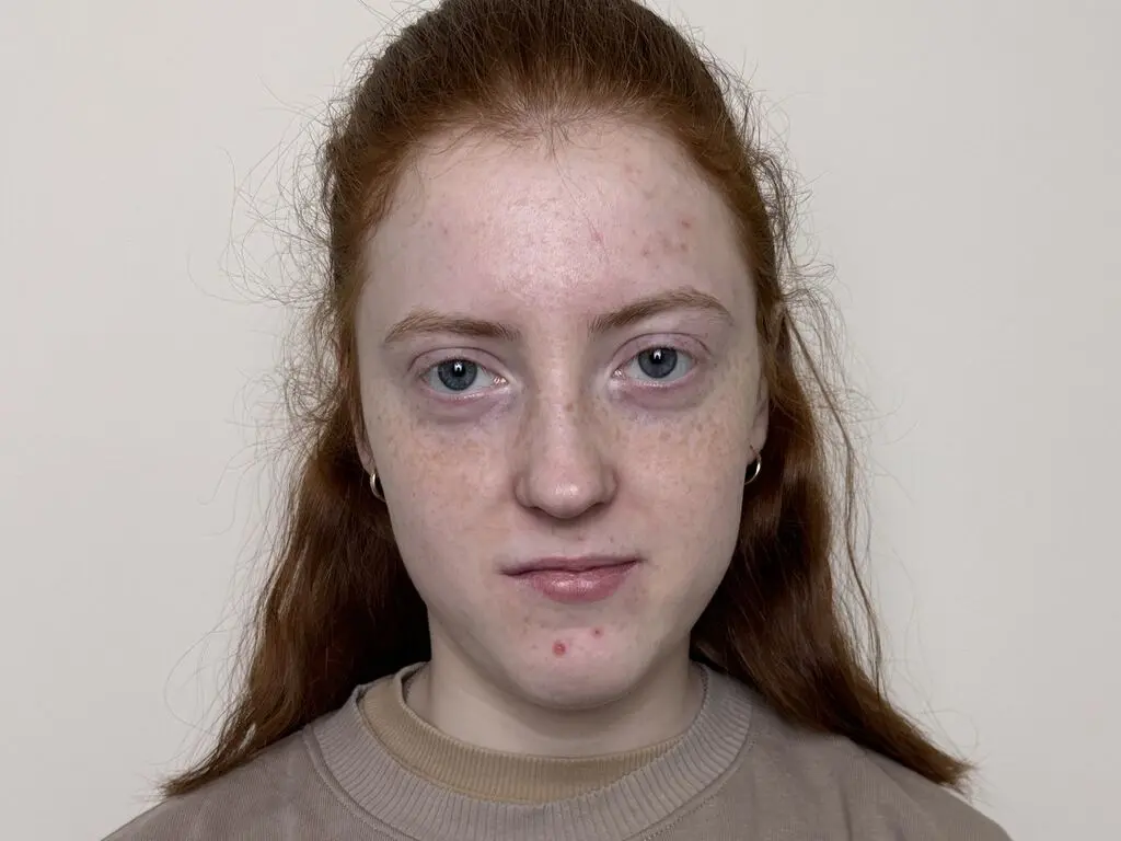 Close-up portrait of a young woman with red hair and freckles wearing a beige sweatshirt against a plain wall.