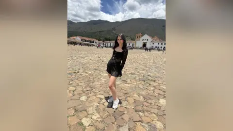 Young woman in a black dress and white sneakers standing on a cobblestone plaza in a scenic mountain village.
