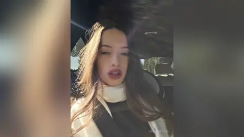 Young woman with long brown hair wearing a white turtleneck sitting in a car during golden hour.