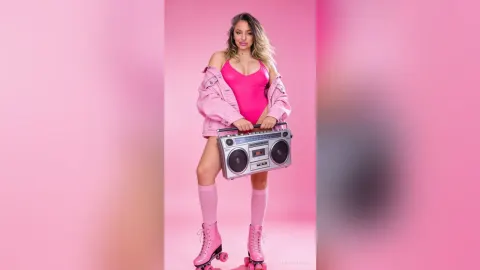 DorothyLake — DorothyLake poses in a pink bodysuit and denim jacket while holding a silver boombox against a pink background.