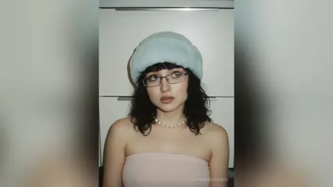 Young woman with dark hair wearing a light blue faux fur hat and pearl necklace in a soft, dreamy indoor setting.