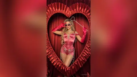 Glamorous woman in a pink corset and red gloves posing inside a heart-shaped frame against red curtains.