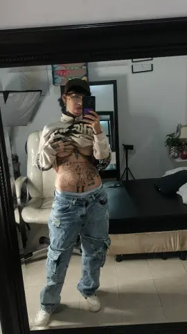 lucirosee poses in a mirror wearing a tan zip-up hoodie and baggy denim cargo pants, showing her stomach and tattoos.