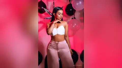 Young woman in a white crop top and pink trousers posing against pink balloons for a celebratory mood.